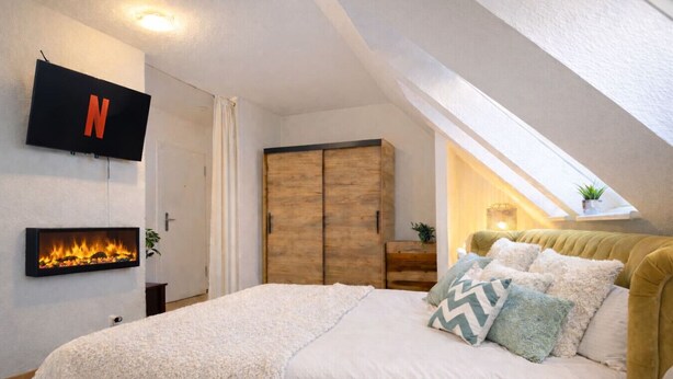 Een slaapkamer met een aan de muur bevestigde televisie, een open haard, een bed met kussens en een houten garderobe.