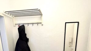 Une petite pièce propre, avec un plancher en bois, une veste noire suspendue à un porte-manteau, un miroir au mur et une petite poubelle.