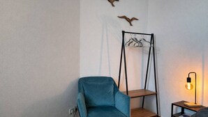 Une pièce avec une chaise bleue, une étagère en bois, un portant à vêtements et une table de chevet avec une lampe.