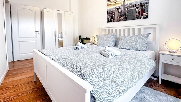 A modern bedroom with a cityscape painting, a neatly made bed, and a bedside table with a lamp. - N&N: Apartment - Schloss & Stadtnähe (Schwerin)