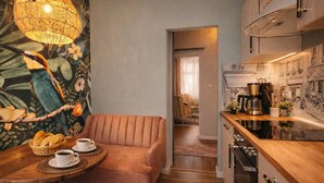 Un salon confortable avec un canapé, une table basse sur laquelle se trouvent des tasses et du pain, une lampe en osier et une cuisine équipée d’une cuisinière et d’une machine à café.