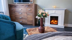 Ein Wohnzimmer mit blauem Sofa, einem Holztisch mit Blumenvase, einer Holzkommode und einem Kamin mit sichtbarem Feuer.