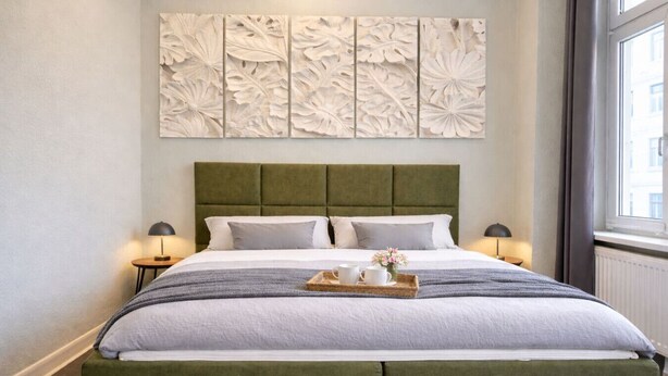 Ein ordentlich bezogenes Bett mit einem grünen Stoffüberzug am Kopfteil, einer dekorativen Wandverkleidung darüber und einem Tablett mit Tassen und Blumen auf dem Bett.