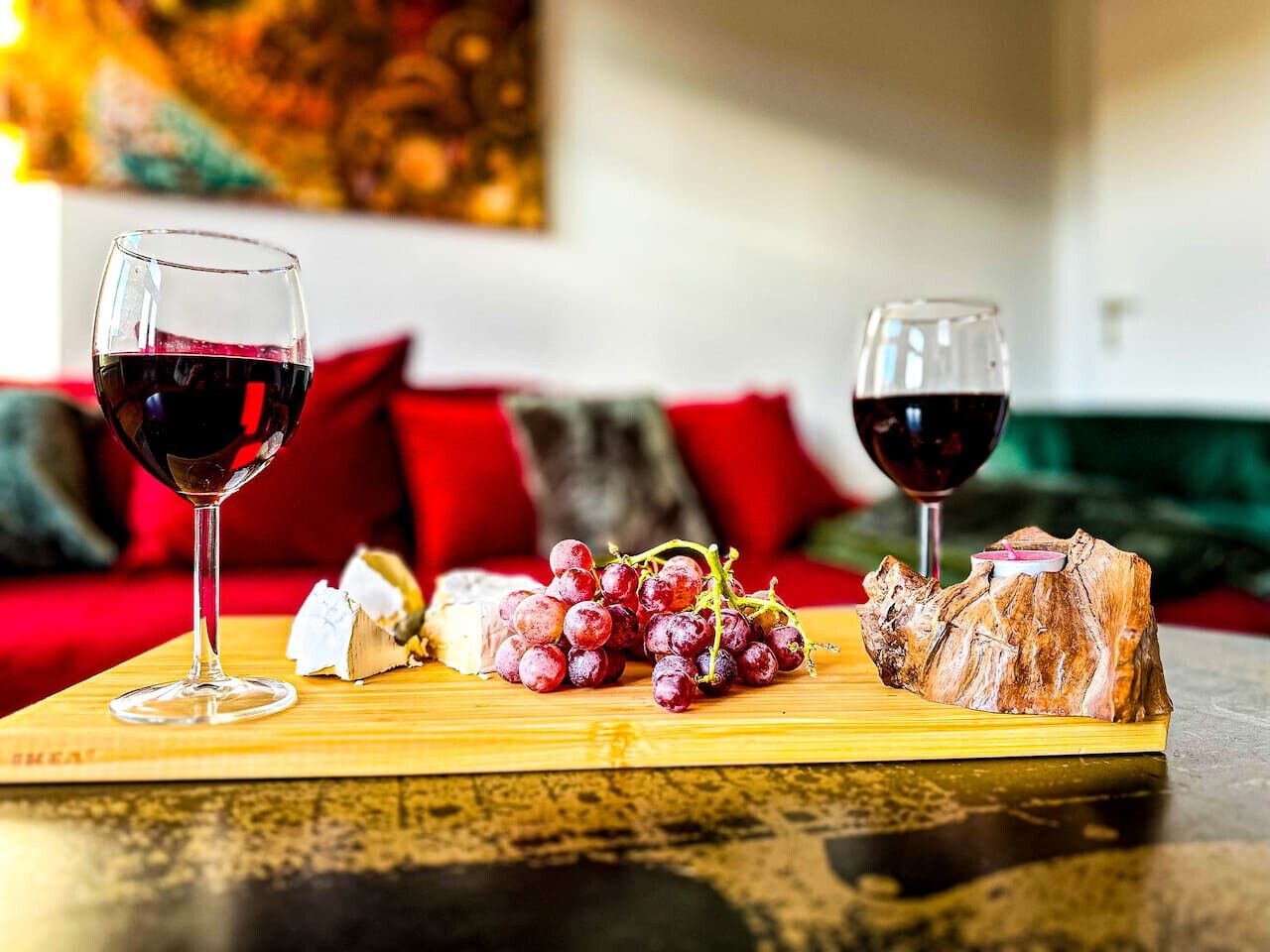A wooden board with a glass of red wine, a piece of cheese, and a bunch of grapes on a table.