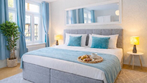 Een keurig opgemaakte slaapkamer met een bed, twee bedlampen, een nachtkastje, een potplant en een raam met gordijnen.
