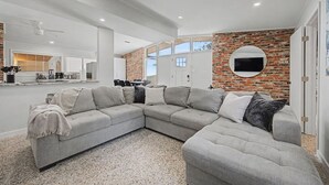 A modern living room with a sectional sofa, a kitchen area, and a brick accent wall. - Pool + Hot Tub Retreat · Sleeps 18  · Near Destin (Fort Walton Beach)