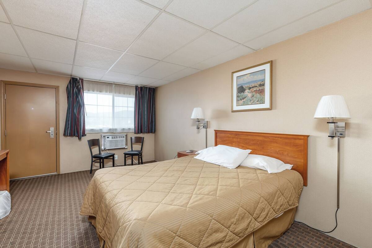 A hotel room with a bed, two bedside lamps, a chair, a desk, an air conditioning unit, and a window with curtains.