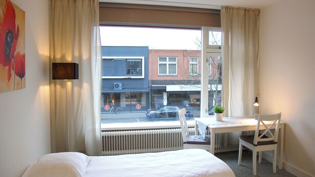 A hotel room with a bed, a desk with a chair, a window with curtains, and a radiator. - Peaceful 1-bedroom studio in charming Enschede perfect for Short-Stay (Enschede)