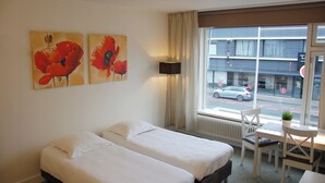 A hotel room with two beds, a window with a view of the street, a table with a chair, and two paintings on the wall. - Peaceful 1-bedroom studio in charming Enschede perfect for Short-Stay (Enschede)