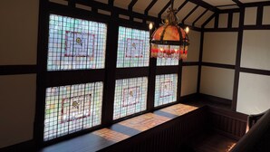 Une pièce dotée d’un plafond haut et de poutres apparentes, agrémentée d’une grande fenêtre à vitraux aux motifs géométriques et d’un lustre suspendu.