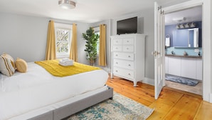 A bedroom with a bed, a bedside plant, a white dresser, a television, and an open door to a bathroom.