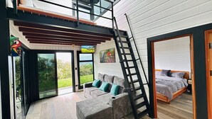 A modern interior with a loft bedroom, a staircase, a sofa with cushions, and a large window offering a view of greenery.