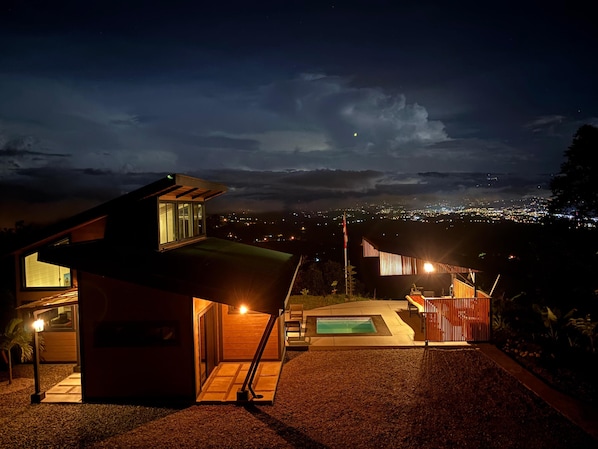 A modern house with a swimming pool, a patio, and a view of the city at night.