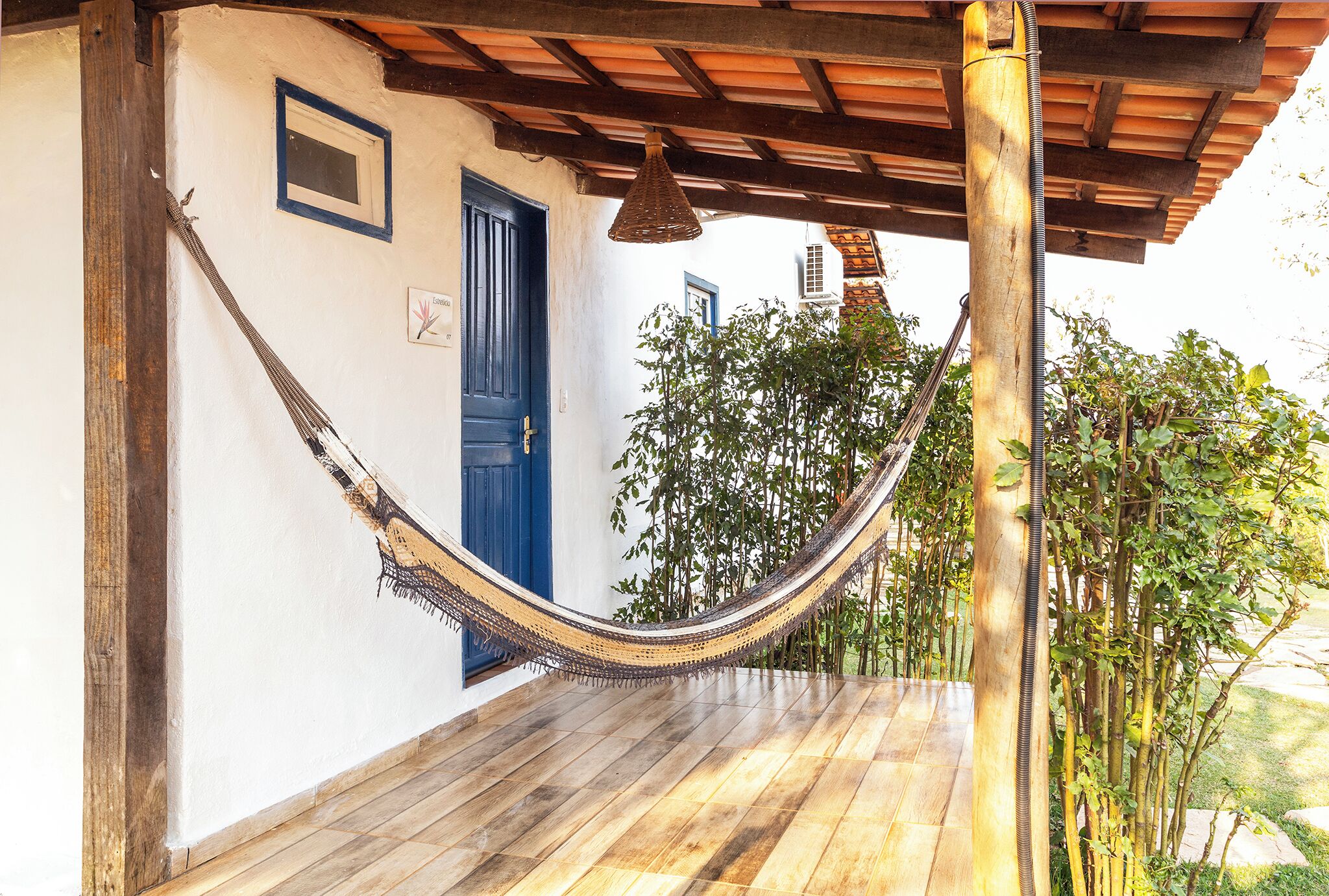 A hammock under a wooden pergola, a blue door, and a white wall with a small window.