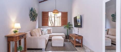 A living room with a sofa, ottoman, wooden side tables, a lamp, a TV, and a large mirror.