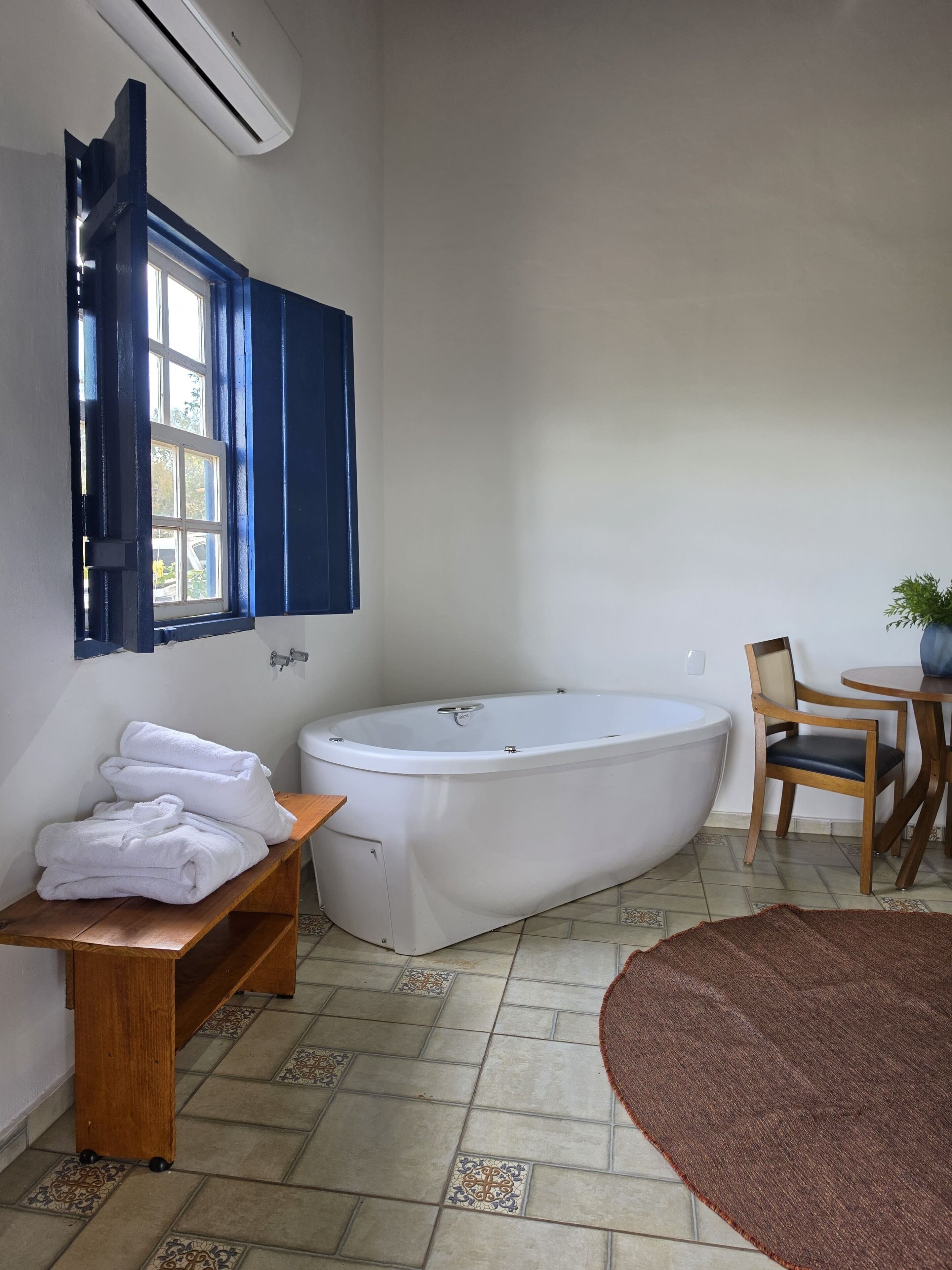 Un baño con una bañera, un banco de madera, una silla, una mesita con una planta y baldosas de suelo estampadas.