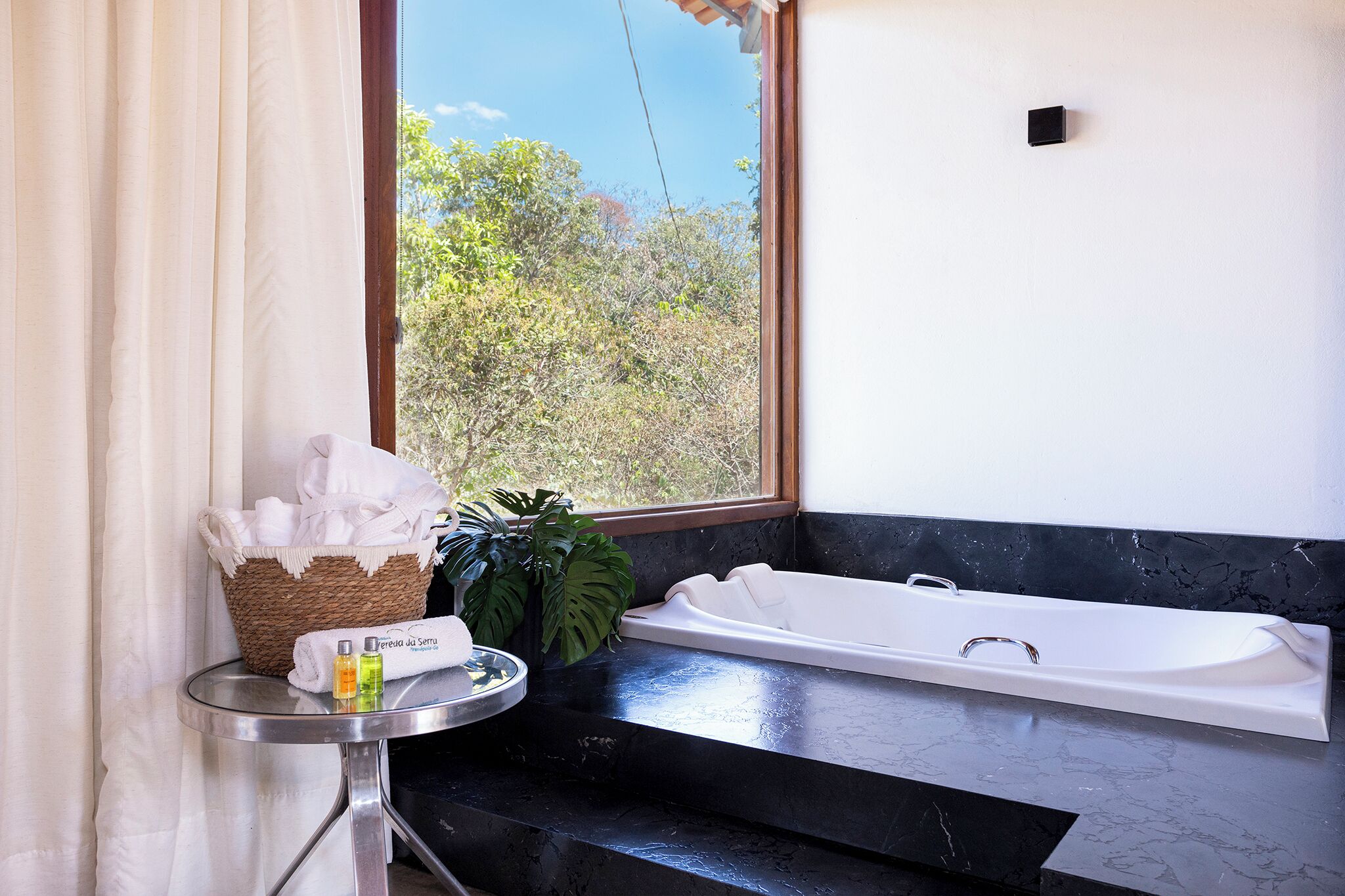 A bathroom with a bathtub, a small table with toiletries, and a view of trees outside.