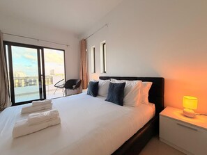 A hotel room with a bed, a chair, a nightstand, and a view of the outside.