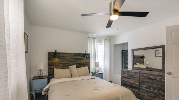 A bedroom with a bed, a nightstand, a dresser, a ceiling fan, and a window with curtains. - Work, Relax & Explore The 'Burgh! McKeesport Home (McKeesport)