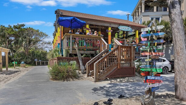 Un edificio frente a la playa con una terraza de madera, un poste con indicaciones y una zona de arena con zapatos.