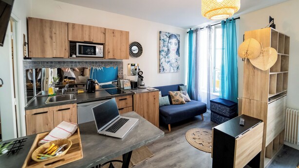 Una cocina moderna con armarios de madera, un microondas y un lavamanos. Hay una mesa de comedor con una computadora portátil y una cesta de utensilios. Un sofá y una mesa de centro están en la zona de estar, con un ventanal y cortinas.