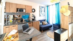 Un espacio habitable compacto con kitchenette, un comedor con una computadora portátil y una zona de estar con sofá y cortinas.