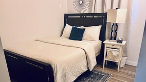 Ein Bett mit Kopfteil, ein Nachttisch mit Lampe, ein kleines Regal und ein Teppich auf Holzdielen.