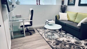 Ein modernes Wohnzimmer mit einem Glastisch, einem schwarzen Bürostuhl, einem runden Couchtisch, einer dunkelgrauen Couch mit grünen und weißen Kissen und einem gemusterten Teppich.