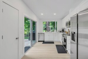 Eine moderne Küche mit weißen Schränken, einem Edelstahlkühlschrank und Blick auf einen Wald durch ein großes Fenster.