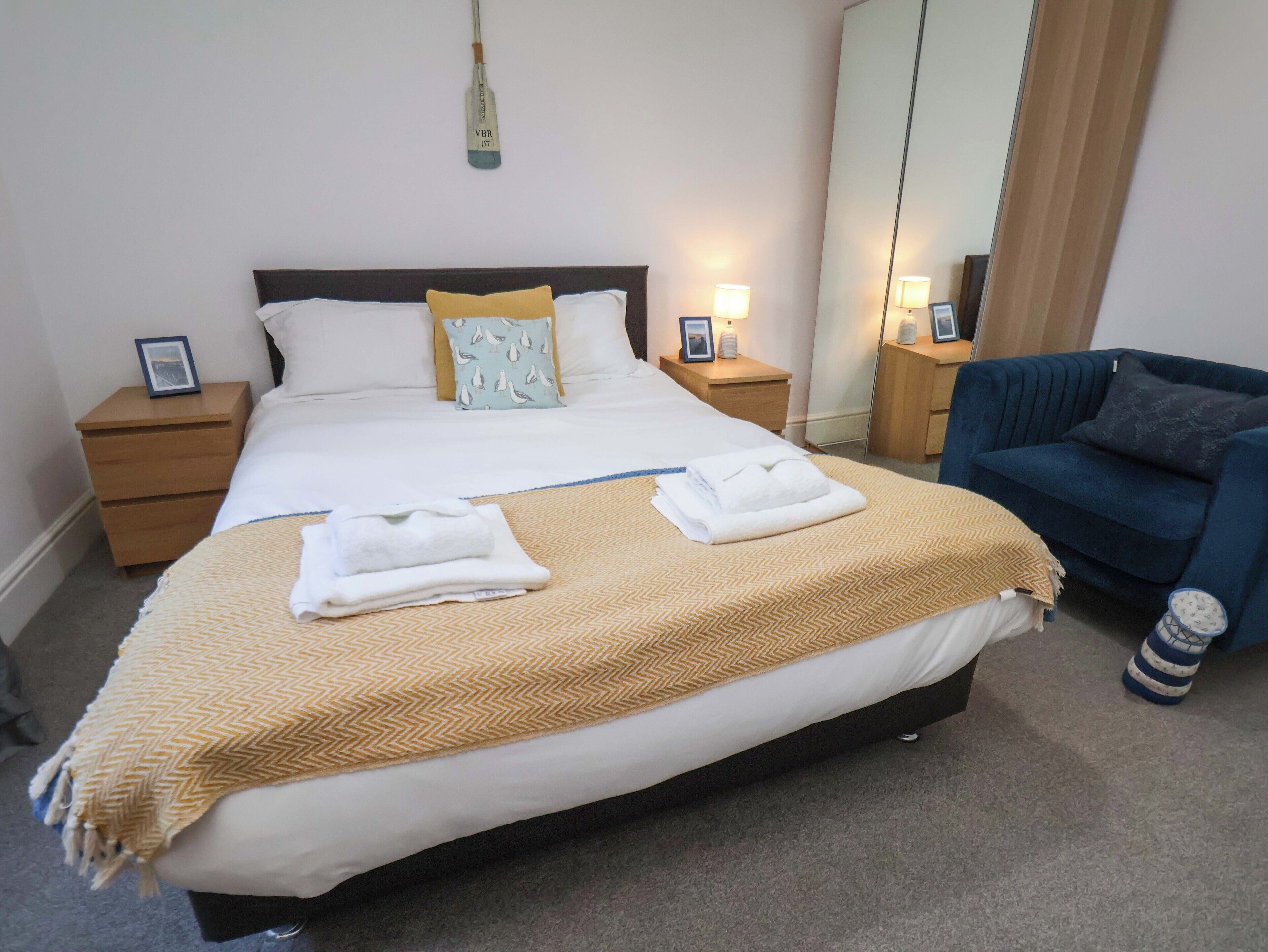 A neatly made bed with two folded towels, a bedside table with a lamp, a mirror, and a blue armchair.