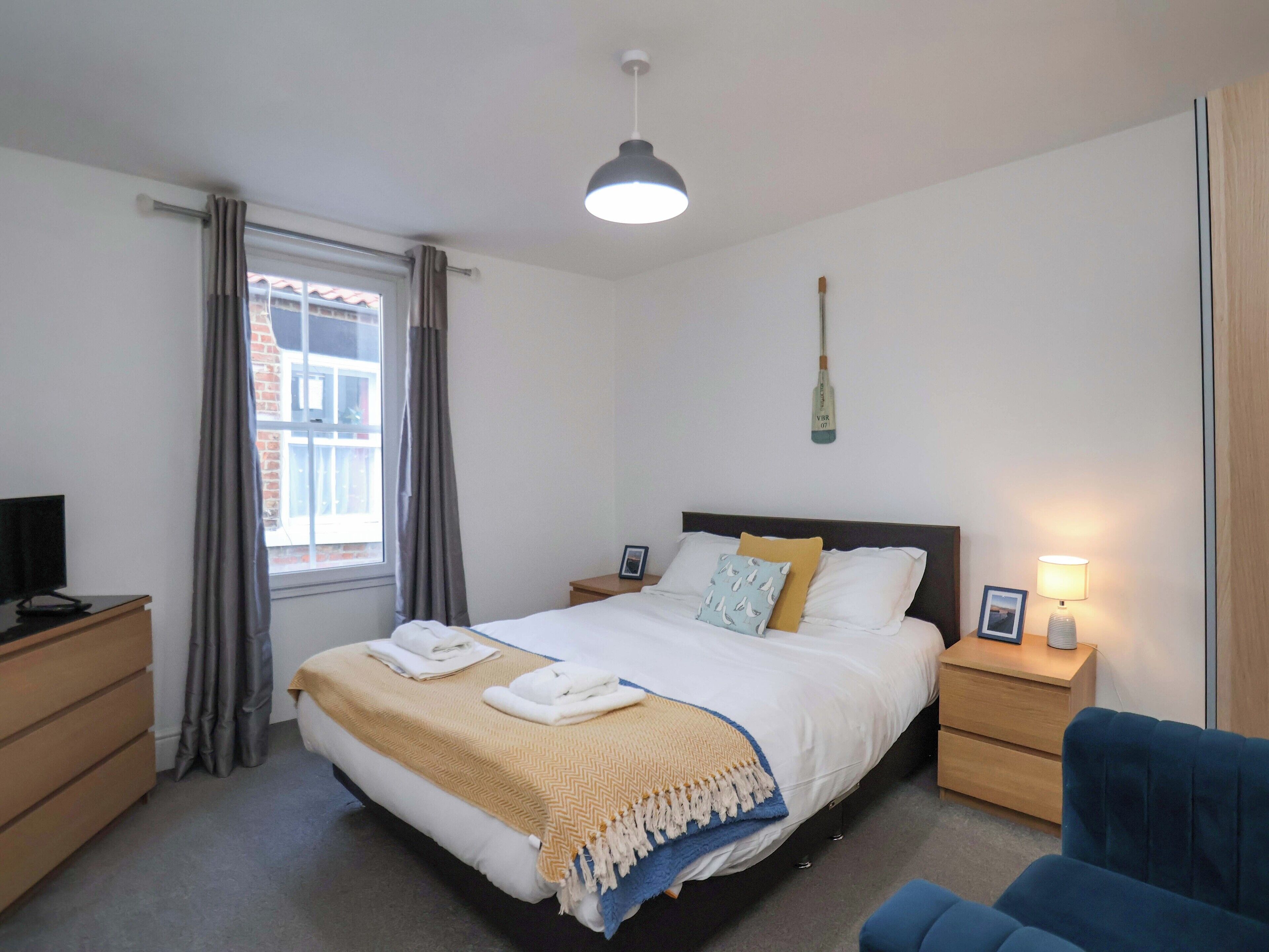 A bedroom with a bed, bedside tables, a desk, a chair, a window with curtains, and a hanging lamp.