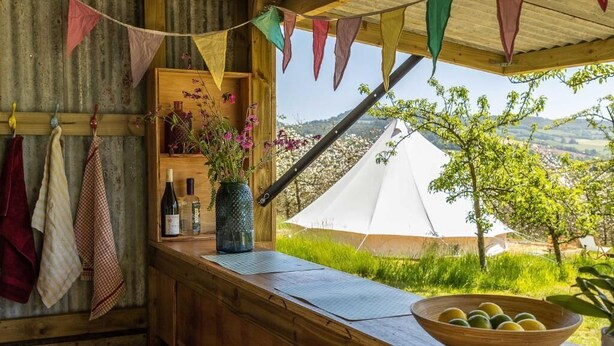 Ein rustikales Holzhaus mit einer Holzbank, einem Holzregal mit Flaschen und einer Blumenvase, sowie Blick auf ein weißes Zelt und viel Grün.