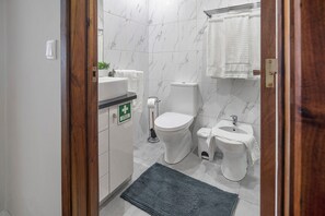 A bathroom with a toilet, bidet, sink, and a green emergency exit sign. - Apartment 'Sérénité' with Balcony, Wi-Fi and Air Conditioning (Ponta Delgada)
