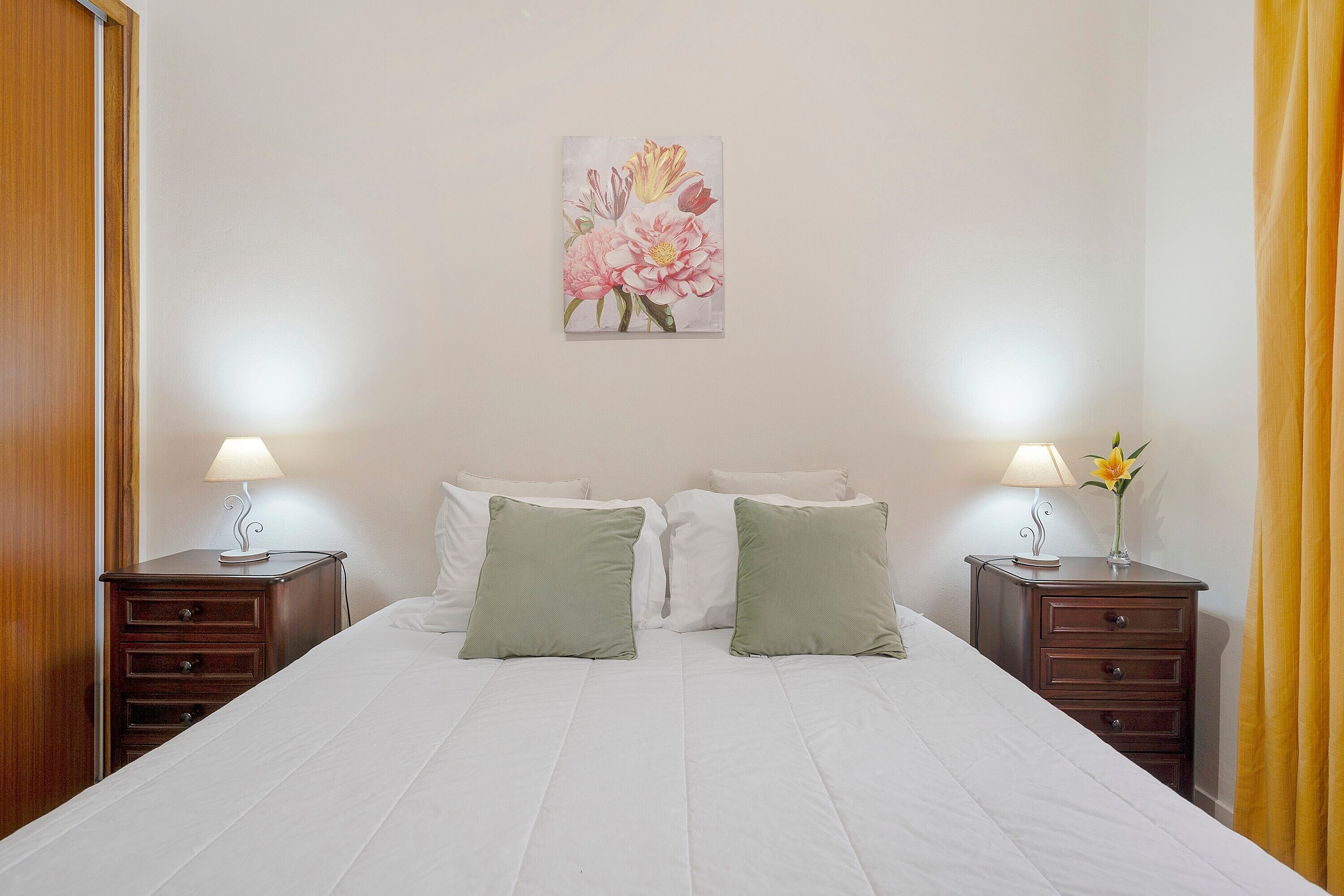 A neatly made bed with white bedding, two bedside tables with lamps, and a floral artwork on the wall.