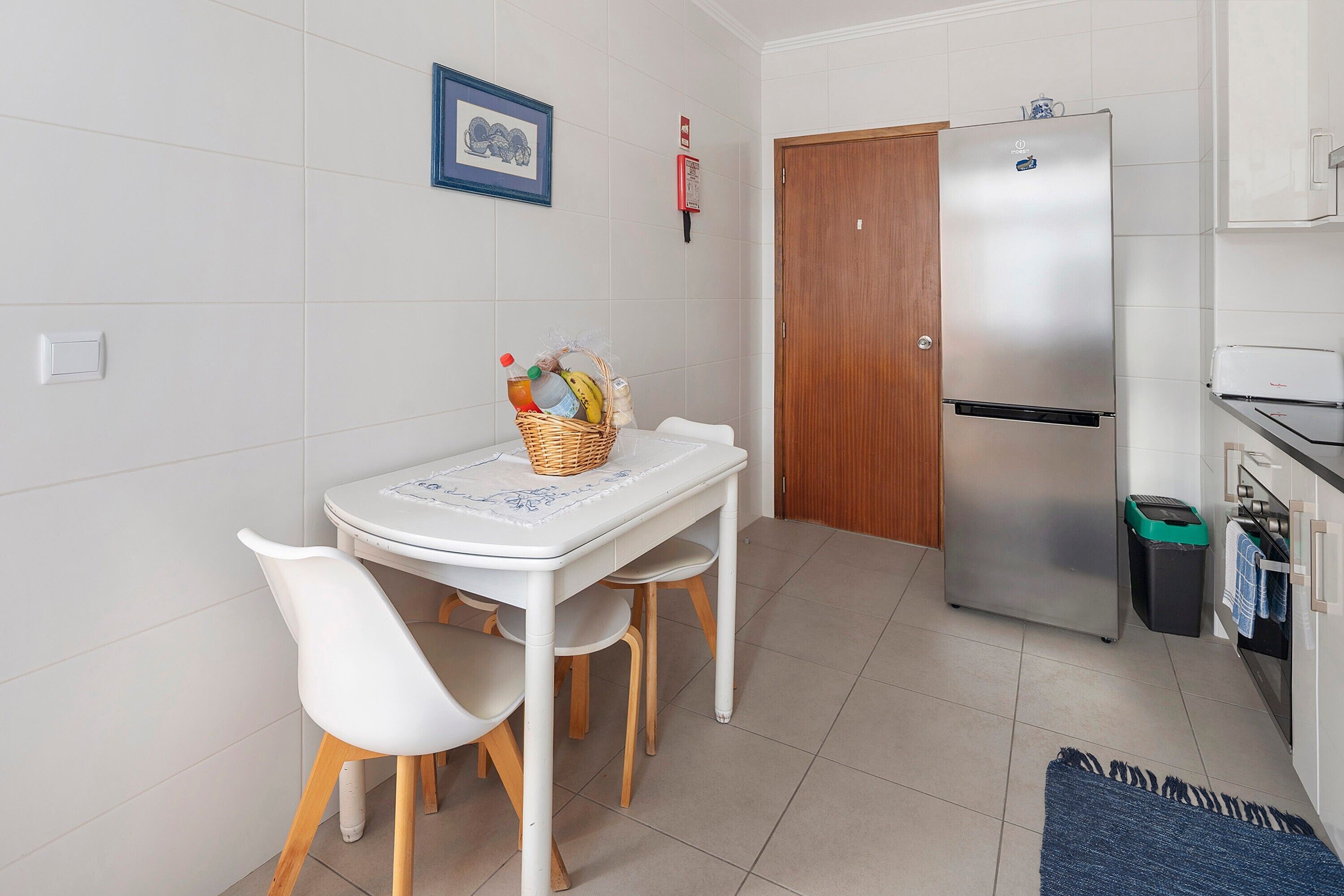 A kitchen with a white table, two white chairs, a stainless steel refrigerator, a wooden door, and a tiled floor.