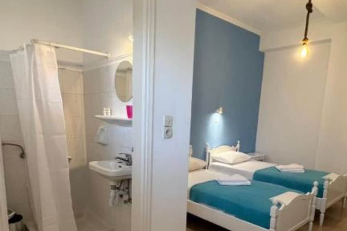 A hotel room with a shower, a sink, two beds with blue bedding, and a light fixture on the ceiling.