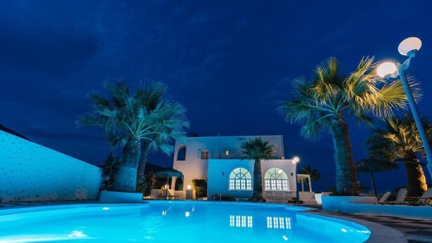 A pool with a building in the background, palm trees, and a street lamp. - Blue Life hotel (Santorini)