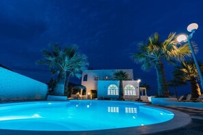 A pool with a building in the background, palm trees, and a street lamp. - Blue Life hotel (Santorini)