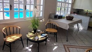 A spacious room with large arched windows overlooking a pool, a round table with chairs, and a kitchen area. - Blue Life hotel (Santorini)