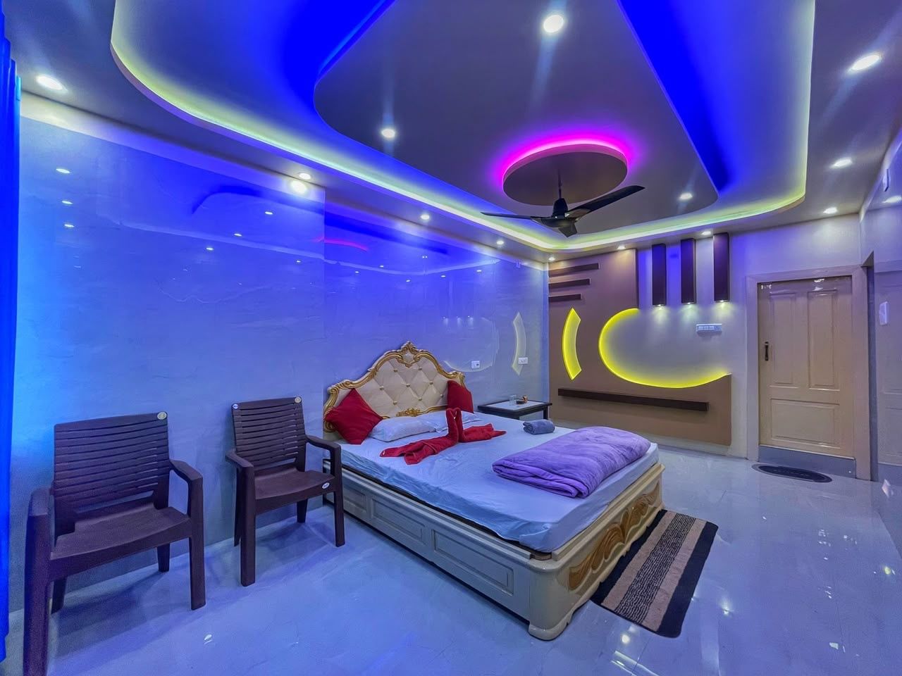 A modern bedroom with a bed, two chairs, a ceiling fan, and a wall-mounted light fixture.