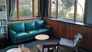 Een kamer met een groene leren bank, een klein rond tafeltje, een houten stoel, een boekenkast en grote ramen met uitzicht op groen.