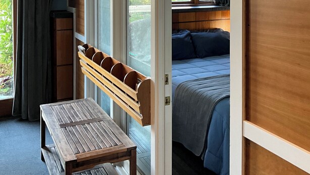 Een kamer met een houten bank, een bed met een blauwe plaid, een raam met gordijnen en een brandblusser.