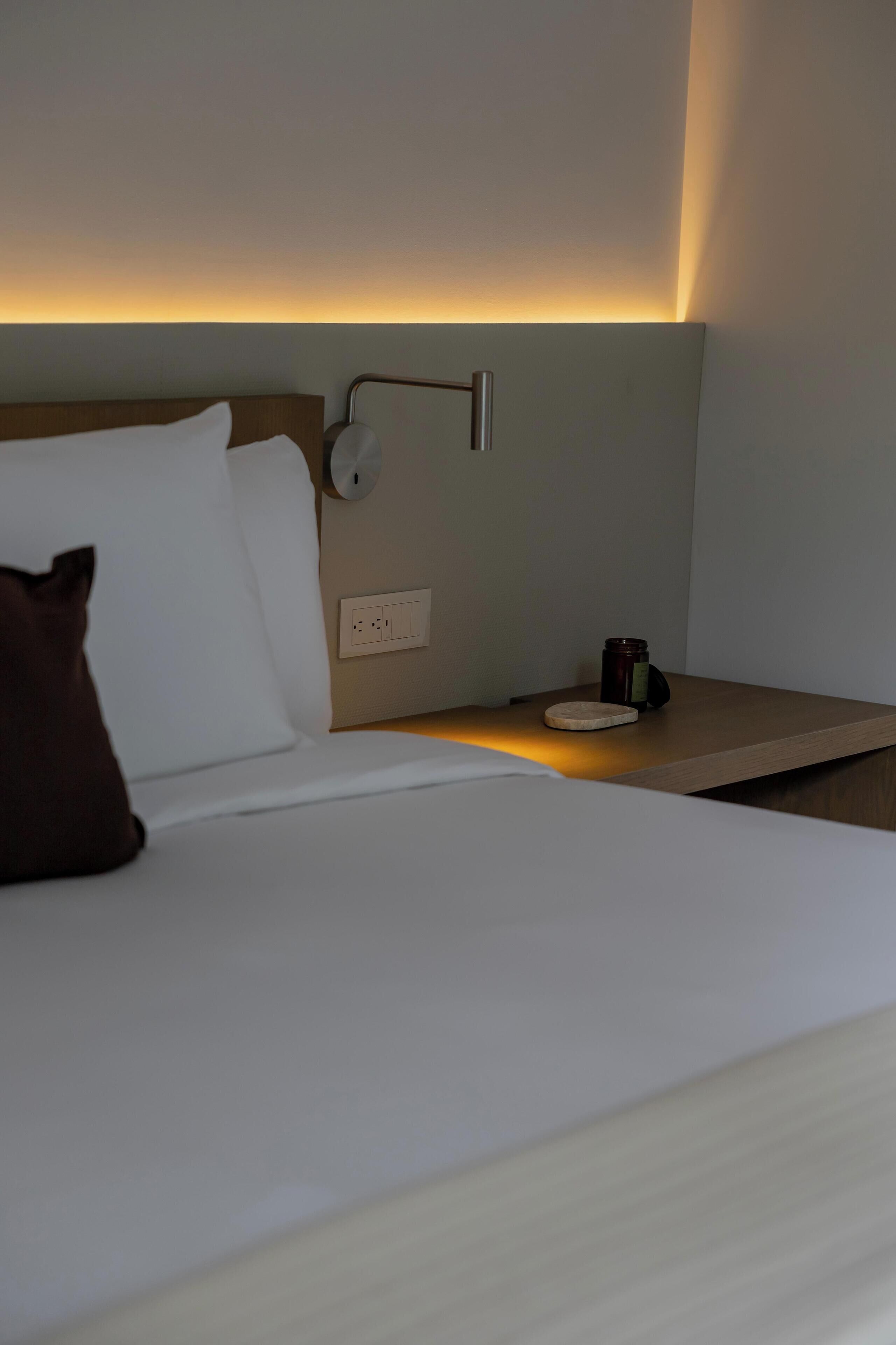 A hotel room with a neatly made bed, a bedside table with a lamp, and a power outlet.