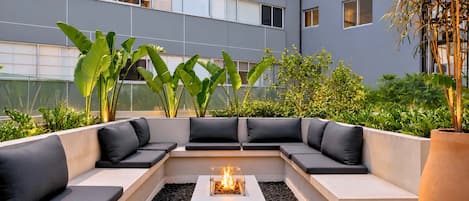 A modern outdoor seating area with a fire pit, surrounded by plants and a building in the background.
