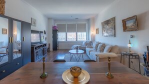 A modern living room with a dining area, large windows, and a view of the city. - Master King plus 2 Queens by NYC & onsite gym (Jersey City)