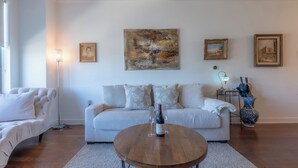 A living room with a sofa, a coffee table with a bottle and glasses, a floor lamp, and framed artwork on the wall. - Master King plus 2 Queens by NYC & onsite gym (Jersey City)