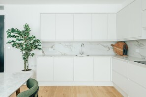 A modern kitchen with white cabinetry, a marble backsplash, and a wooden dining table with a green chair. - Luxury 2BR Apartment in Mayfair (London)
