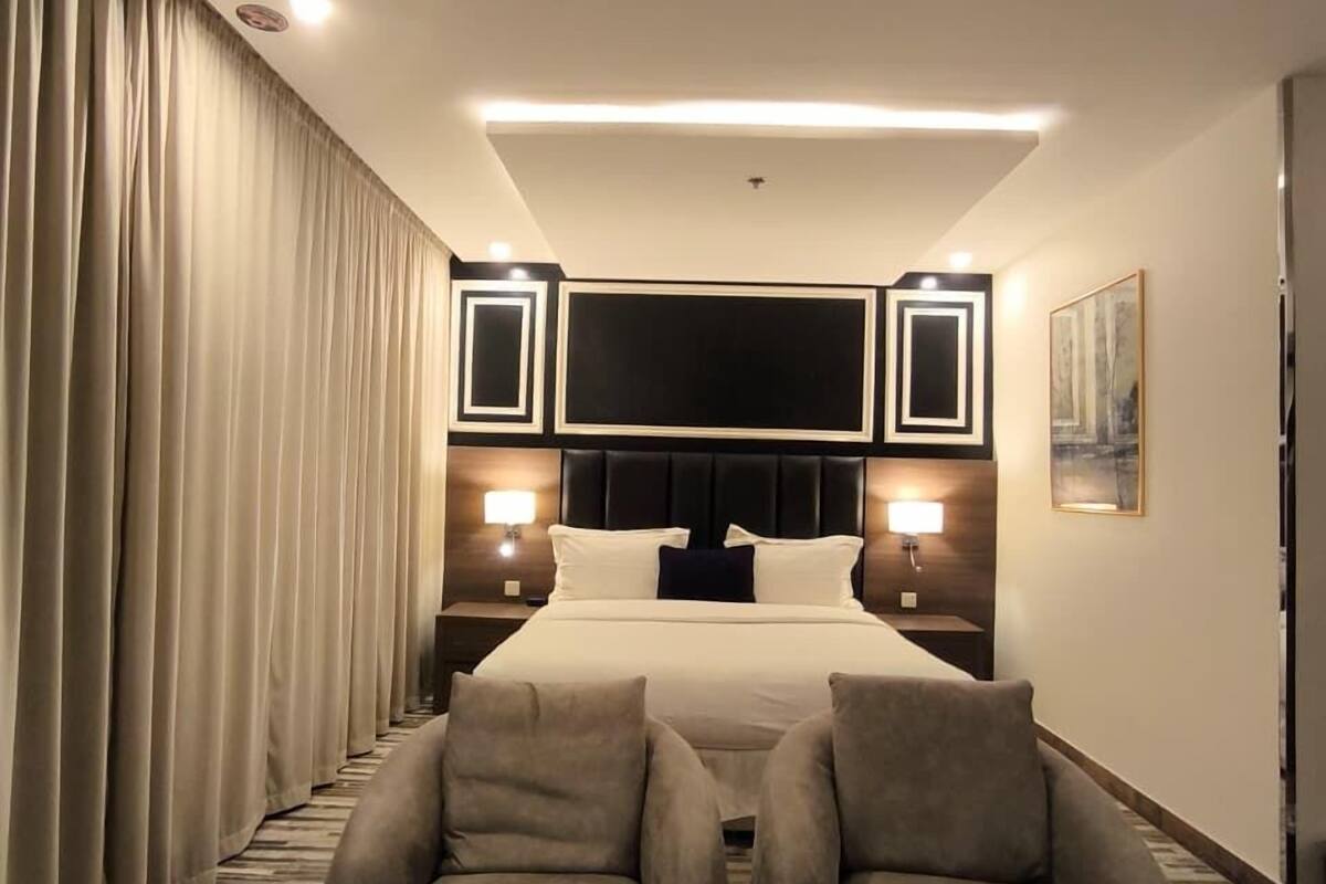 A hotel bedroom with a bed, two armchairs, a small table, a TV, and a painting on the wall.