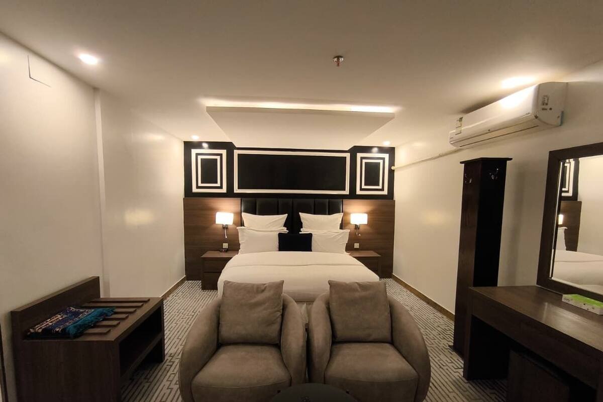 A hotel room with a bed, bedside tables, a sofa, a small table, a mirror, and an air conditioning unit.