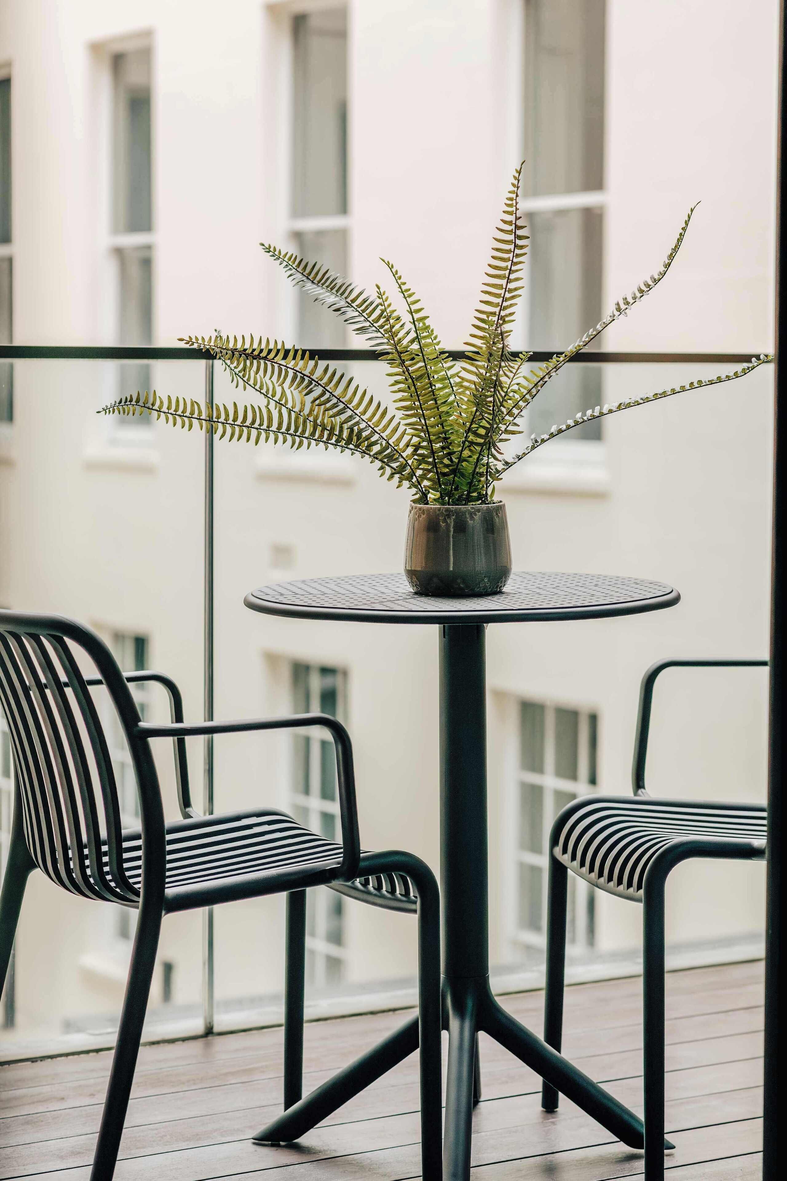 A balcony with a round table and two chairs, a potted fern, and a building in the background.